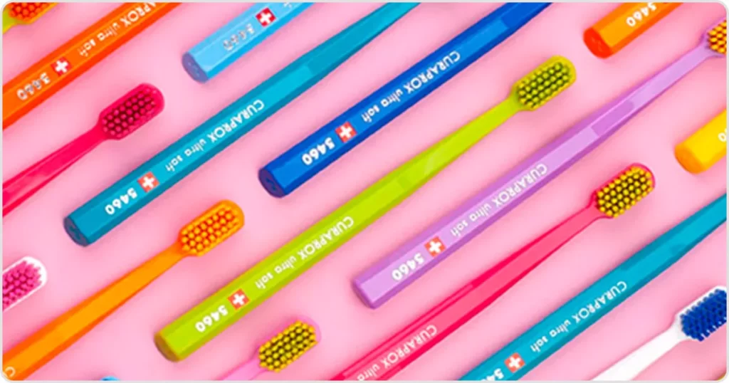 Top view of Curaprox toothbrushes in various colors on pink background. Product available at Hola Sonríe, dentist in Coyoacán, CDMX.